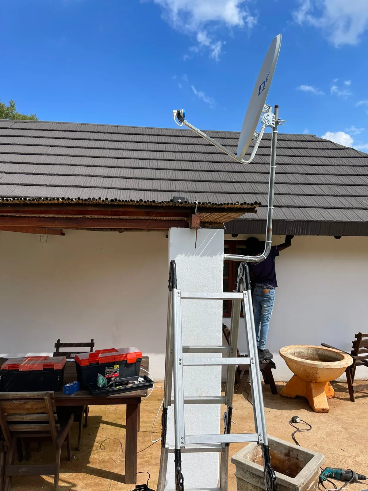 DSTV technician on a ladder mounting a dish on a tiled roof, toolbox on the ground